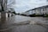 Floodwaters cover a residential street, partially submerging garages and houses on either side. Trees without leaves are visible, and the sky is overcast, adding to the somber atmosphere.