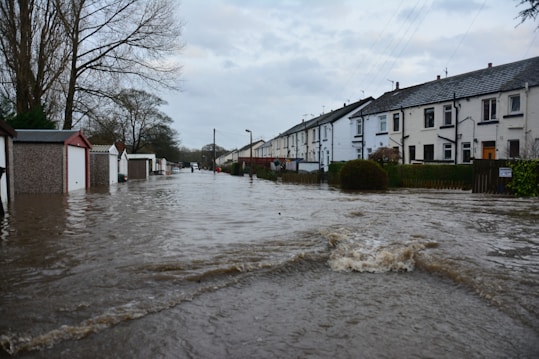 Floodwaters cover a residential street, partially submerging garages and houses on either side. Trees without leaves are visible, and the sky is overcast, adding to the somber atmosphere.