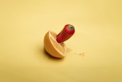 A red chili pepper is inserted into a sliced orange half, both positioned on a smooth yellow background. The composition blends elements of fruit and spice in a visually striking manner.