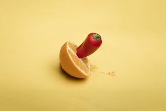 A red chili pepper is inserted into a sliced orange half, both positioned on a smooth yellow background. The composition blends elements of fruit and spice in a visually striking manner.