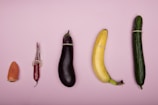 A row of five different fruits and vegetables lined up against a soft pink background. Each item is covered with a condom. From left to right, there is an orange bell pepper, a red chili pepper, an eggplant, a banana, and a cucumber.