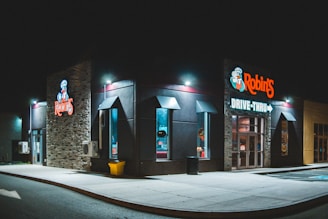 A drive-thru fast food restaurant is illuminated at night, featuring a brightly lit sign with modern architecture. The building has stone and dark paneling, with large windows and small outdoor lights along the roofline. The atmosphere is calm and quiet, with no visible activity around.