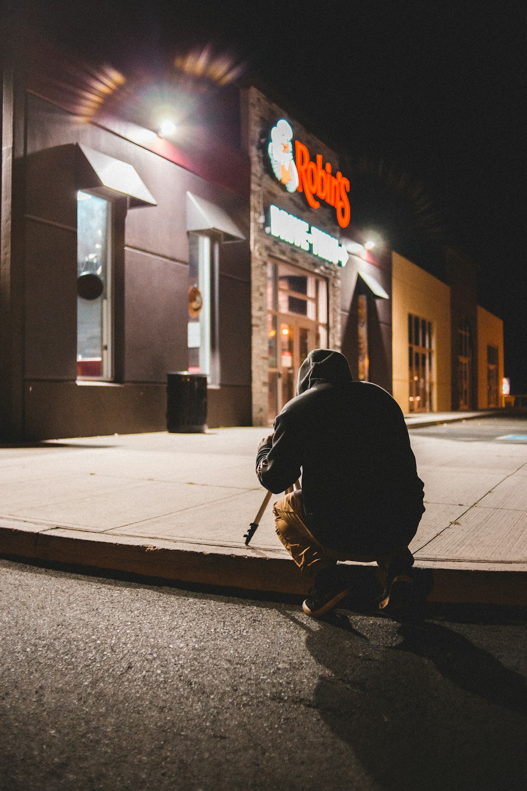 man in black hoodie and black pants sitting on sidewalk during night time