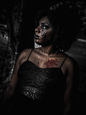 A woman with dark curly hair gazes upward, wearing a textured black dress and a necklace. Her face and chest are marked with blood and special effects makeup, creating a horror-themed appearance. The background is dark and shadowy, enhancing the eerie and haunting atmosphere.