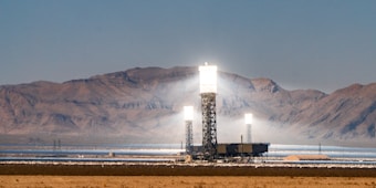 Three tall, industrial-looking solar power towers are located in a desert landscape with mountains in the background. The towers are brightly illuminated at the top, likely from concentrated sunlight. Surrounding the towers are arrays of mirrors used to direct sunlight towards the towers.
