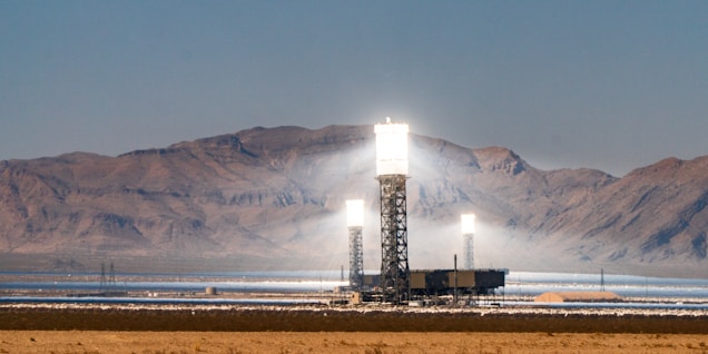 Three tall, industrial-looking solar power towers are located in a desert landscape with mountains in the background. The towers are brightly illuminated at the top, likely from concentrated sunlight. Surrounding the towers are arrays of mirrors used to direct sunlight towards the towers.