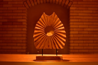 A wooden sculpture with a central circular cut-out stands on a wooden platform. The sculpture's surface is shaped with radiating lines that create a pattern. Behind it lies a brick wall with an arched pattern at the top, softly illuminated by warm lighting.