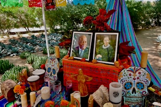 An altar decorated with vibrant, colorful patterns and objects, possibly for the Day of the Dead celebration. It includes framed photos of two individuals, intricately decorated sugar skulls, marigold flowers, candles, and decorative crafts. Papel picado, a traditional paper decoration, hangs above in bright colors. The backdrop includes lush green plants and a striped fabric in blue and purple tones.