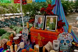 An altar decorated with vibrant, colorful patterns and objects, possibly for the Day of the Dead celebration. It includes framed photos of two individuals, intricately decorated sugar skulls, marigold flowers, candles, and decorative crafts. Papel picado, a traditional paper decoration, hangs above in bright colors. The backdrop includes lush green plants and a striped fabric in blue and purple tones.