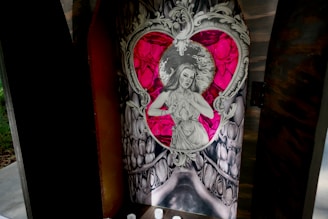 An intricately designed artwork featuring a grayscale drawing of a woman framed within a heart shape. Surrounding the heart is an elaborate, ornate pattern reminiscent of baroque art. The background includes vibrant shades of red and pink, providing a stark contrast to the monochromatic figure. The setting appears to be enclosed, like a shrine or a niche, suggesting a spiritual or reverential context.
