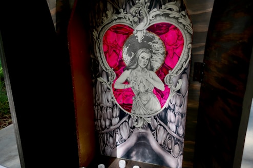 An intricately designed artwork featuring a grayscale drawing of a woman framed within a heart shape. Surrounding the heart is an elaborate, ornate pattern reminiscent of baroque art. The background includes vibrant shades of red and pink, providing a stark contrast to the monochromatic figure. The setting appears to be enclosed, like a shrine or a niche, suggesting a spiritual or reverential context.