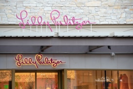 The image features a storefront with the name 'Lilly Pulitzer' displayed prominently in pink signage. The upper part of the store facade is composed of light-colored bricks, creating a stylish and clean appearance. Below, the glass display window showcases mannequins dressed in colorful clothing, adding a vibrant touch to the scene.