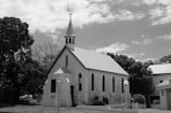 A small church building with a steeple and cross is situated among trees under a partly cloudy sky. The architecture includes arched windows and a simple entrance. The scene is in black and white, adding a timeless quality to the image.