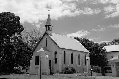 A small church building with a steeple and cross is situated among trees under a partly cloudy sky. The architecture includes arched windows and a simple entrance. The scene is in black and white, adding a timeless quality to the image.