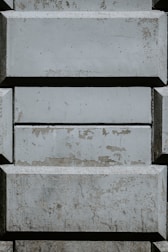 A pattern of rectangular concrete blocks arranged in a staggered, brick-like fashion. The surface of the blocks shows signs of wear and aging, with patches of peeling paint and discoloration. Shadows cast from the edges add depth to the textured surface.