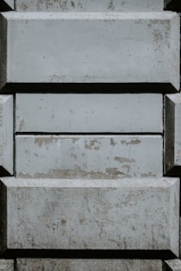 A pattern of rectangular concrete blocks arranged in a staggered, brick-like fashion. The surface of the blocks shows signs of wear and aging, with patches of peeling paint and discoloration. Shadows cast from the edges add depth to the textured surface.
