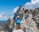 Smiling woman wearing climbing gear, standing on a rocky cliff with a bright blue sky behind her.