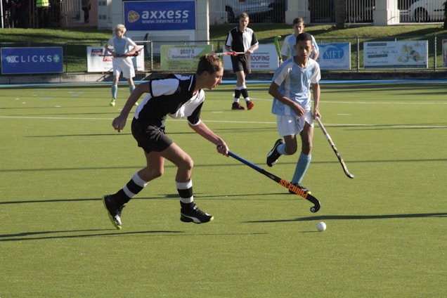 Two young players on a field hockey pitch are engaged in an intense game. One player in a black and white uniform is in control of the ball, while another in a light blue and white uniform attempts to intercept. The background features advertisements and other players watching the game.