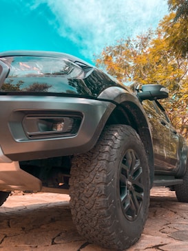 A rugged vehicle is photographed from a low angle, emphasizing its large, textured front tire and sleek body. The vehicle is parked on a stone-paved surface under a bright blue sky. Surrounding the car are lush trees with green and golden leaves, suggesting a sunny day.