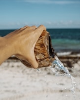 A hand is holding a coconut shell, pouring clear water out onto a sandy beach with a view of the ocean in the background. The sunlight creates a sparkling effect on the water droplets.