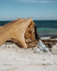 A hand is holding a coconut shell, pouring clear water out onto a sandy beach with a view of the ocean in the background. The sunlight creates a sparkling effect on the water droplets.