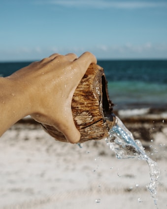 A hand is holding a coconut shell, pouring clear water out onto a sandy beach with a view of the ocean in the background. The sunlight creates a sparkling effect on the water droplets.
