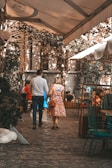 A couple is walking hand in hand through a charming outdoor café setting. The scene is shaded by a canopy with dappled sunlight filtering through. The cobblestone path and surrounding greenery create a cozy and intimate ambiance. The man wears a patterned shirt and dark pants, while the woman is in a colorful floral dress. Additional café tables and chairs can be seen in the background.