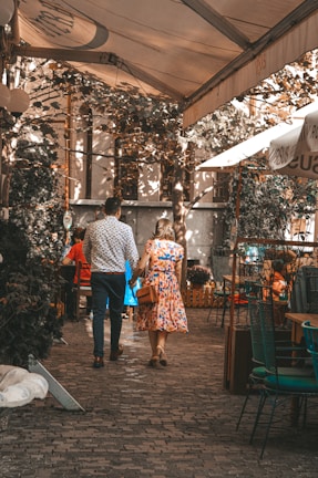 A couple is walking hand in hand through a charming outdoor café setting. The scene is shaded by a canopy with dappled sunlight filtering through. The cobblestone path and surrounding greenery create a cozy and intimate ambiance. The man wears a patterned shirt and dark pants, while the woman is in a colorful floral dress. Additional café tables and chairs can be seen in the background.