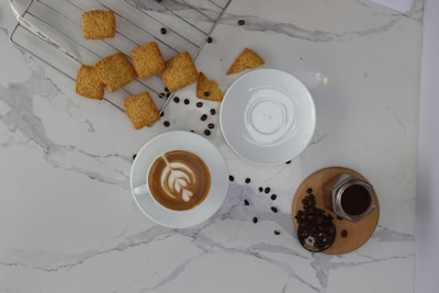 A marble countertop holds a cup of latte with intricate latte art, a small glass jar filled with coffee, and scattered coffee beans. A wire rack displays several square-shaped cookies, some of which have fallen onto the countertop. An empty white saucer is placed beside the coffee cup.