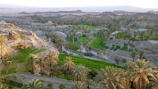 A panoramic view of a lush oasis landscape featuring green fields interspersed with numerous palm trees. The surrounding arid mountains provide a stark contrast to the vibrant greenery below, highlighting the fertility of the area. A winding road cuts through the scene, adding a sense of depth and leading the eye through the oasis. The lighting suggests a time of day close to sunset, casting warm tones across the landscape.