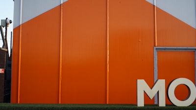 A large industrial building with bright orange and gray panels, featuring the partial letters 'MO' in white at the bottom right. A security camera is mounted on the side of an adjacent structure.