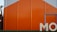 A large industrial building with bright orange and gray panels, featuring the partial letters 'MO' in white at the bottom right. A security camera is mounted on the side of an adjacent structure.