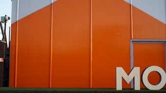 A large industrial building with bright orange and gray panels, featuring the partial letters 'MO' in white at the bottom right. A security camera is mounted on the side of an adjacent structure.