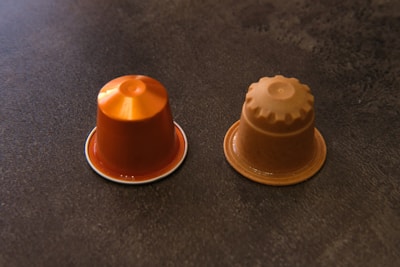 Two coffee capsules placed side by side on a dark textured surface. The capsule on the left is shiny and orange, while the capsule on the right is matte and brown with a ridged top.
