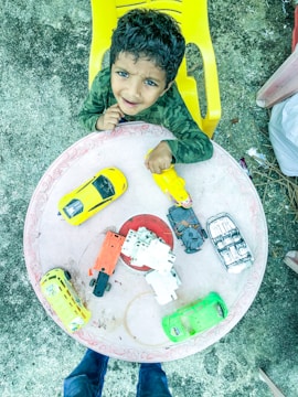 A child with curly hair, wearing a green camouflage-patterned shirt, is seated on a yellow chair. In front of the child is a round table with several toy cars of various colors, including yellow, orange, and green. The child is looking up with a cheerful expression.