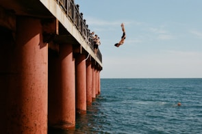 A person is diving off a pier into the blue ocean while wearing swim trunks. The pier is supported by large, rust-colored pillars. Several onlookers are standing on the pier, watching the diver. The water reflects the clear sky above.