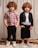 Two young children with curly hair are standing side by side against a light-colored curtain backdrop. The child on the left is wearing a red, white, and blue plaid shirt with black pants and white sneakers. The child on the right is dressed in a light blue shirt, black jacket, black shorts, and blue shoes. Both children have their hands in their pockets and are looking towards the camera.