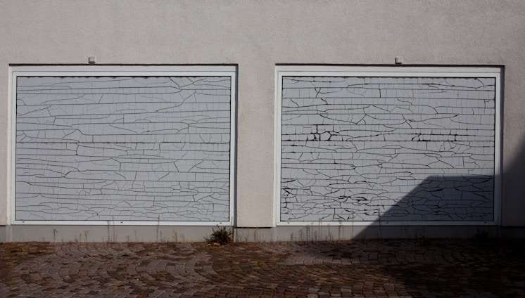 Two white garage doors with a cracked pattern covering their surface, situated on a gray wall. The ground consists of cobblestones with some grass growing between the gaps. A shadow is cast on the right side of the image.