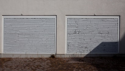 Two white garage doors with a cracked pattern covering their surface, situated on a gray wall. The ground consists of cobblestones with some grass growing between the gaps. A shadow is cast on the right side of the image.