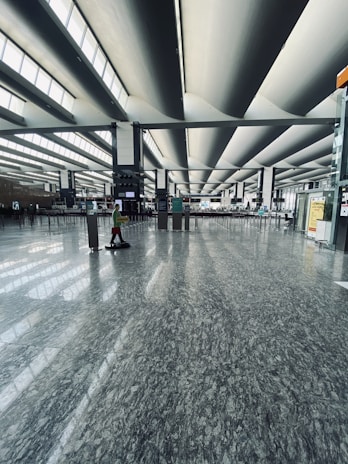 A spacious modern airport terminal with large architectural ceiling design and wide open spaces. Natural light floods through the long horizontal windows, casting patterns on the polished marble floor. Minimal foot traffic is visible, with a lone person walking with a wheeled suitcase.