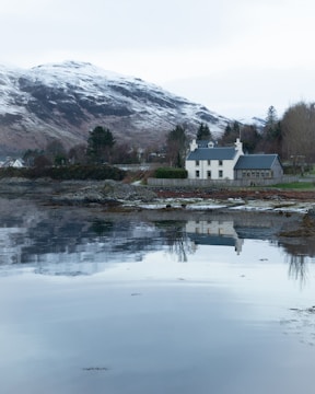 A serene landscape with a house by the water's edge, surrounded by trees. Snow-covered mountains rise majestically in the background, and the calm water reflects the scenery, including the house and trees.