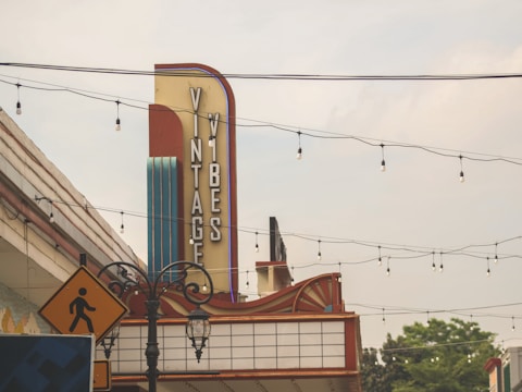 A retro-style sign reading 'Vintage Vibes' is prominently displayed against a blue sky. The sign features a vertical design with bold letters on a colorful background. Strings of hanging lights stretch across the sky, and an ornate streetlamp is visible below. A yellow pedestrian crossing sign is positioned in front of the marquee.