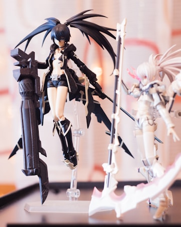 Detailed figures in a display setting, featuring two action figures with elaborate armor and weapons. One has long dark blue hair styled into large ponytails, wearing dark shorts and a coat, with a large weapon. The other figure has light-colored hair and armor, possibly holding a weapon.