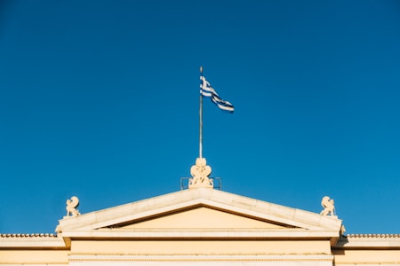A classical building with a triangular pediment and a Greek flag flying on top against a clear blue sky. Ornamental sculptures decorate the corners of the roof.