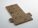 Wooden letter tiles arranged in a crossword style, forming words such as JOY, AWE, AND, MORE, DERIVE, WOVEN, and FINE. Each tile has a letter and a small number indicating its value, similar to Scrabble tiles.