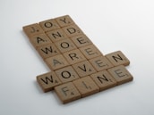 Wooden letter tiles arranged in a crossword style, forming words such as JOY, AWE, AND, MORE, DERIVE, WOVEN, and FINE. Each tile has a letter and a small number indicating its value, similar to Scrabble tiles.