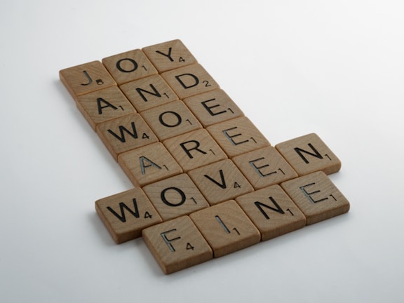 Wooden letter tiles arranged in a crossword style, forming words such as JOY, AWE, AND, MORE, DERIVE, WOVEN, and FINE. Each tile has a letter and a small number indicating its value, similar to Scrabble tiles.
