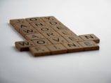Wooden letter tiles arranged in a crossword style, displaying words such as JOY, AWARE, WORD, and WOVEN against a clean, white background.