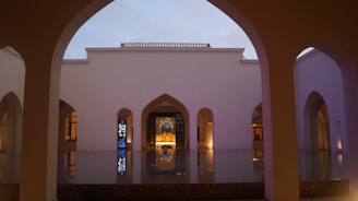 An architectural scene featuring a serene courtyard with symmetrical arches surrounding a reflective water surface. The building's facade is illuminated softly, highlighting its intricate design. The sky appears dim as dusk approaches, adding a serene atmosphere.