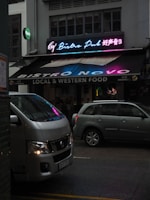 A vibrant bistro pub with a neon sign displaying colorful text in both English and another language. The establishment offers local and western food and advertises live football. Parked in front of the pub are two vehicles, a white van and a silver car, on a dimly lit street.